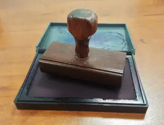 Ilustrasi: Sebuah stempel kayu diletakkan di atas bantalan tinta yang terbuka di meja.