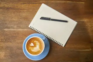 Ilustrasi: Secangkir kopi latte, buku catatan bergaris, dan sebuah pulpen di atas meja kayu.