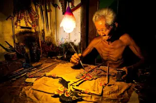 Ilustrasi: Seorang pria lanjut usia sedang melukis wayang kulit di bawah lampu gantung.