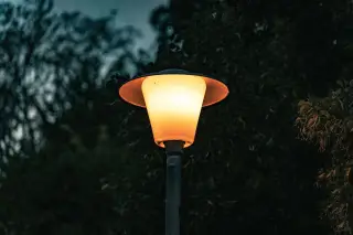 Ilustrasi: Lampu taman atau jalan menyala dengan cahaya hangat di depan dedaunan gelap.