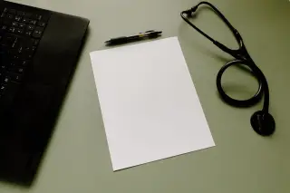 Ilustrasi: Kertas putih kosong, pena, stetoskop hitam, dan sebagian laptop di atas meja.