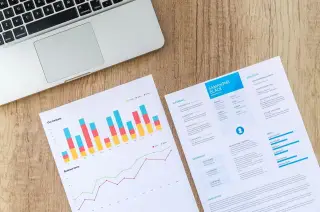 Ilustrasi: Laptop, lembar grafik bisnis, dan dokumen resume di atas meja kayu.