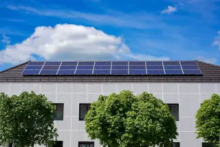 Ilustrasi: Deretan panel surya terpasang di atap gedung berlatar langit biru dan pepohonan hijau.