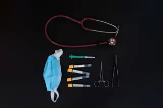 Ilustrasi: Stetoskop, masker medis, jarum suntik, tabung sampel, dan gunting bedah di atas latar hitam.