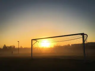 Ilustrasi: Gawang sepak bola di lapangan rumput berkabut dengan latar matahari terbit.