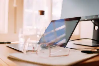 Ilustrasi: Meja kerja dengan laptop terbuka, monitor tambahan, buku catatan, dan kacamata.