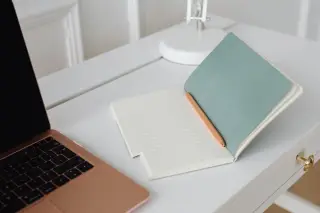 Ilustrasi: Buku catatan terbuka dengan pena di atas meja putih di samping laptop.