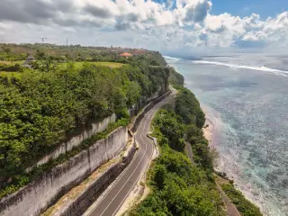 Ilustrasi: Pemandangan udara jalan berliku di tepi tebing dan pantai berkarang dengan laut lepas.