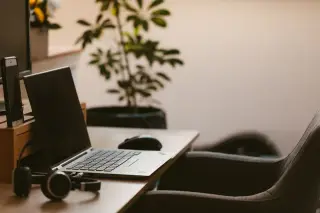 Ilustrasi: Meja kerja dengan laptop terbuka, tetikus, headphone, dan tanaman hias di dalam ruangan.