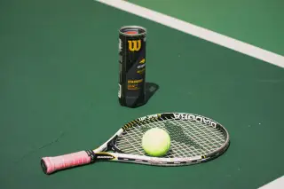 Ilustrasi: Raket tenis, bola tenis, dan wadah bola di atas lapangan tenis hijau bergaris putih.