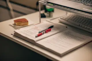 Ilustrasi: Buku terbuka, pulpen, dan laptop di atas meja belajar.