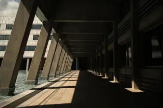 Ilustrasi: Lorong luar gedung bertiang beton dengan bayangan kuat tanpa ada orang.