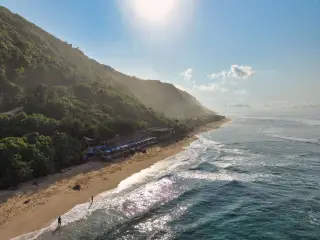 Ilustrasi: Pemandangan udara garis pantai berpasir di bawah tebing hijau dengan sinar matahari terik.