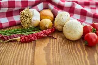 Ilustrasi: Bawang putih, cabai merah kering, jamur, dan tomat di atas meja kayu dengan serbet kotak-kotak.