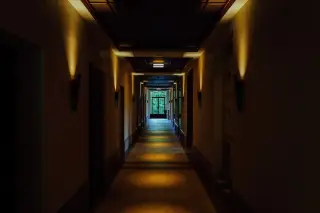 Ilustrasi: Lorong bangunan yang sepi dan remang dengan lampu dinding serta pintu kaca di ujungnya.