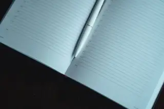 Ilustrasi: Buku agenda terbuka dengan pena putih di atas meja kayu.
