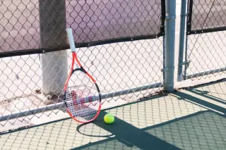 Ilustrasi: Sebuah raket tenis dan bola tergeletak di tepi lapangan tenis di dekat pagar kawat.