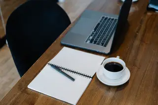 Ilustrasi: Laptop, buku catatan spiral kosong, pulpen, dan secangkir kopi di atas meja kayu.