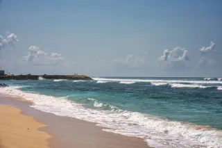 Ilustrasi: Pemandangan pantai berpasir dengan ombak laut biru dan langit cerah berawan.