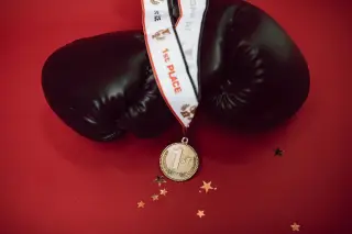 Ilustrasi: Sarung tinju hitam dan medali emas bertuliskan juara pertama di atas latar belakang merah.