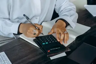 Ilustrasi: Seseorang sedang menulis di buku catatan di dekat kalkulator pada meja kerja.
