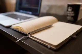 Ilustrasi: Buku catatan terbuka dengan pena di atas meja kerja di samping laptop.