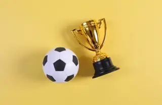 Ilustrasi: Bola sepak mainan dan sebuah piala kecil berwarna emas di atas latar belakang kuning polos.