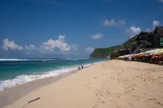 Ilustrasi: Pemandangan pantai berpasir dengan ombak laut, tebing, dan langit biru cerah berawan.