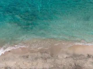 Ilustrasi: Pemandangan dari atas pantai berpasir dengan ombak laut biru kehijauan dan beberapa orang di tepi air.
