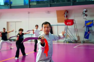 Ilustrasi: Seorang pria dan sekelompok orang berlatih seni bela diri wushu dengan pedang di dalam gedung olahraga.