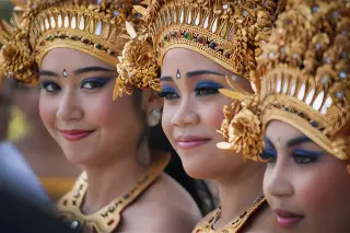 Ilustrasi: Tiga wanita mengenakan riasan dan mahkota tari tradisional Bali berwarna emas.