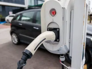 Ilustrasi: Kabel pengisi daya terpasang pada stasiun pengisian kendaraan listrik dengan mobil di latar belakang.