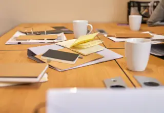 Ilustrasi: Buku catatan, map berkas, dan cangkir putih di atas meja rapat kayu.