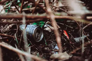 Ilustrasi: Kaleng minuman bekas dan sampah plastik tergeletak di antara semak dan ranting kering.