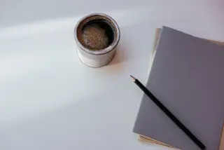 Ilustrasi: Secangkir kopi, pensil hitam, dan buku catatan di atas meja putih.