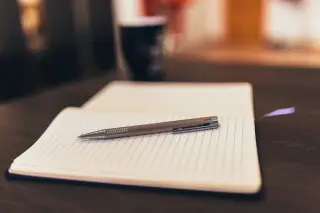 Ilustrasi: Buku catatan bergaris yang terbuka dan pulpen logam di atas meja kayu.