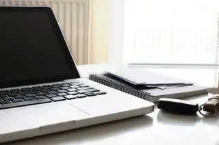 Ilustrasi: Laptop, buku catatan, amplop, dan kunci mobil di atas meja kerja putih.