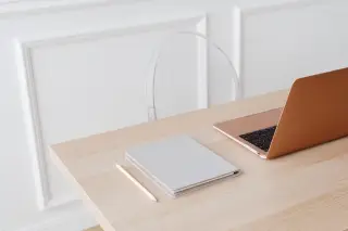 Ilustrasi: Sebuah laptop, buku catatan, dan pensil tertata di atas meja kayu dengan latar dinding putih.